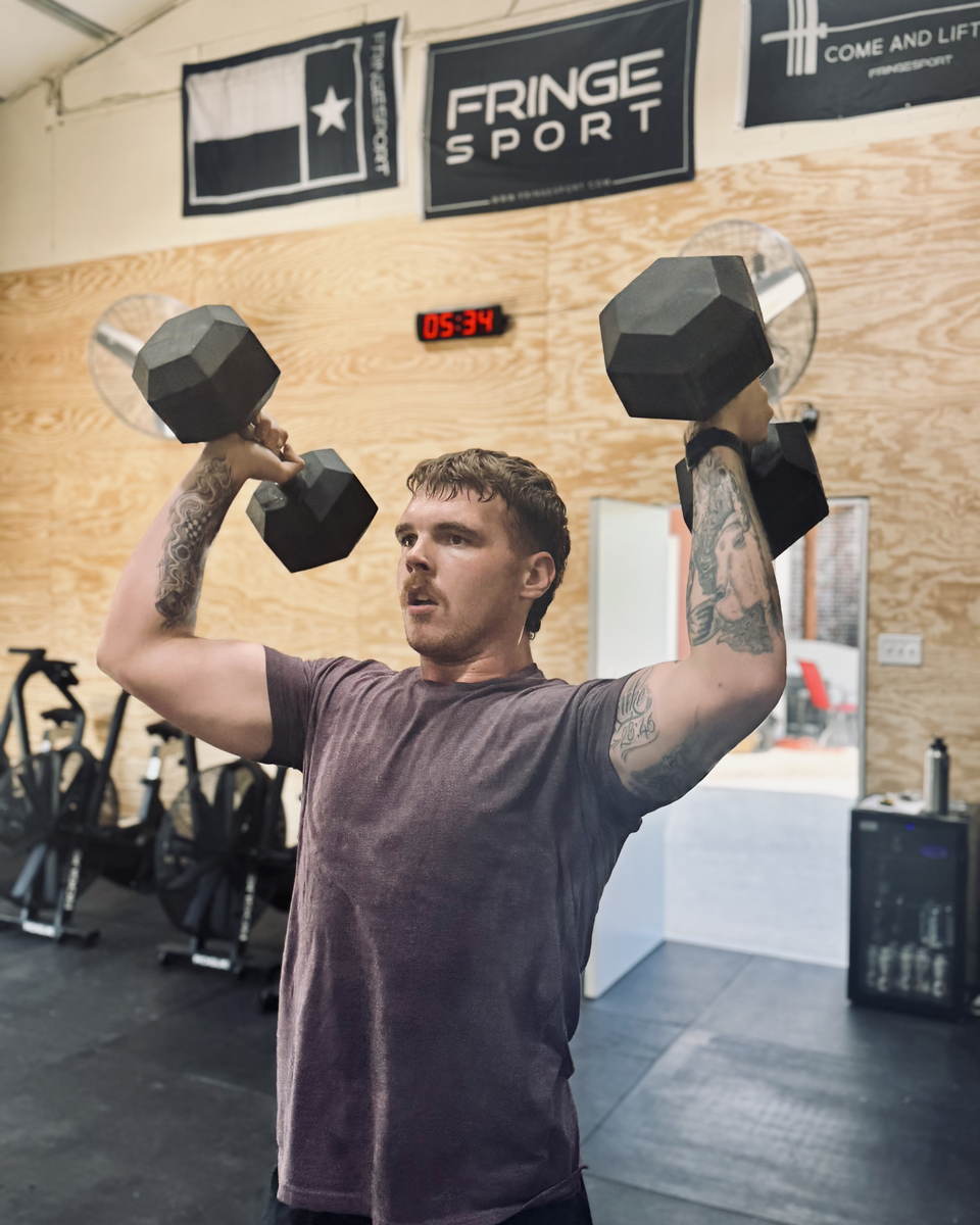A member locking two dumbbells out overhead with the workout clock counting behind him.