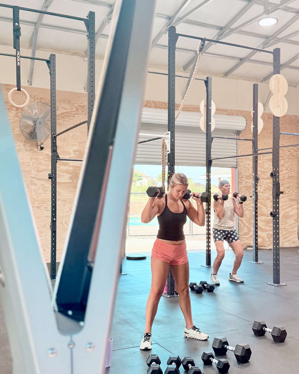 Two members side by side, dumbbells racked at their shoulders, working through a class together in the main training bay.