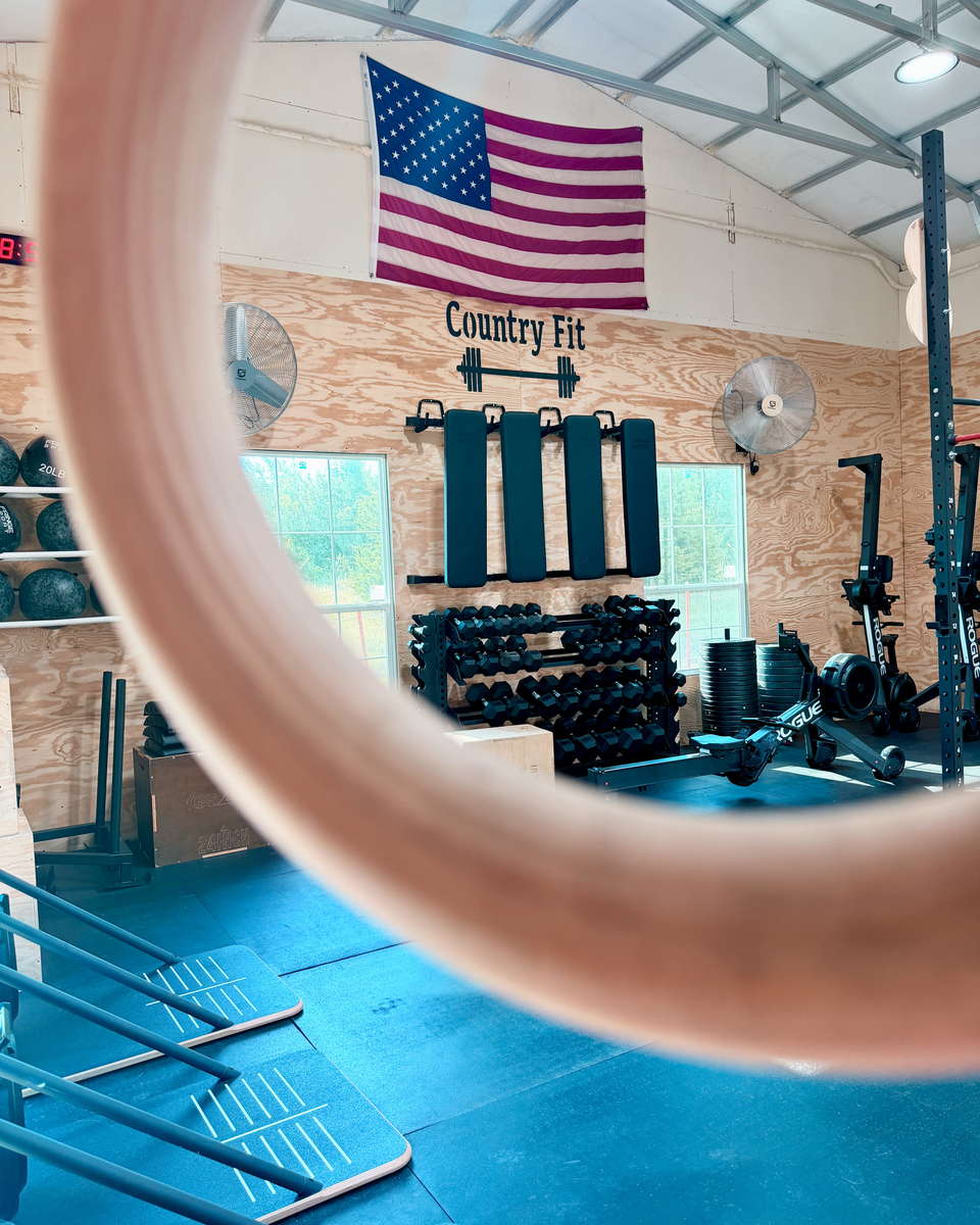 The gym floor framed through a gymnastic ring: dumbbell racks, wall-mounted benches and the Country Fit sign beneath an American flag.