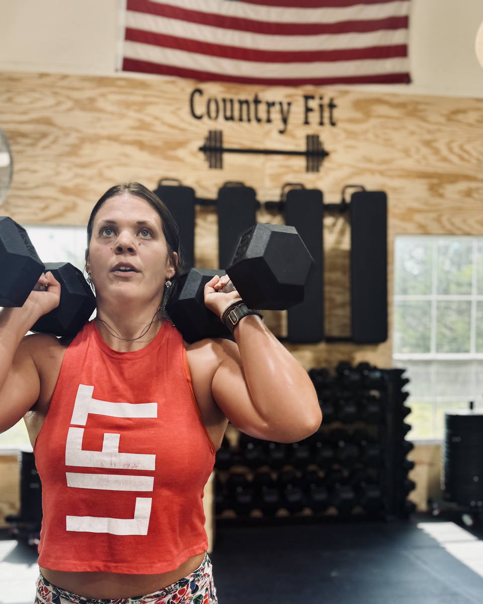 A member holding two dumbbells at her shoulders directly in front of the Country Fit wall sign.