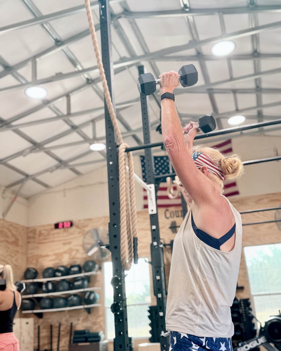 A member pressing a dumbbell overhead next to the climbing rope, wearing a stars-and-stripes headband.