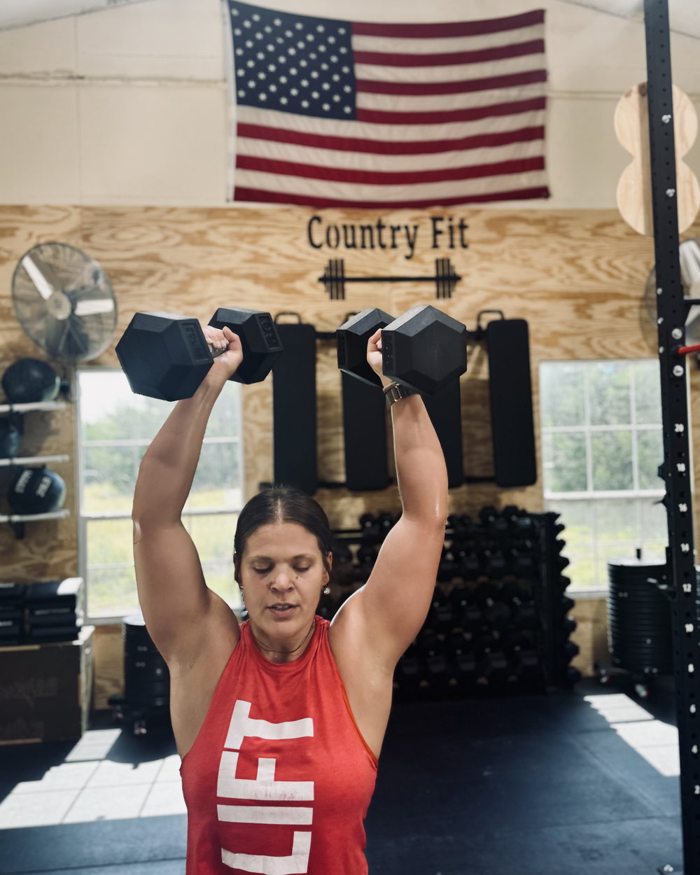 A Country Fit member pressing two dumbbells overhead beneath the gym's hand-painted Country Fit sign and an American flag.