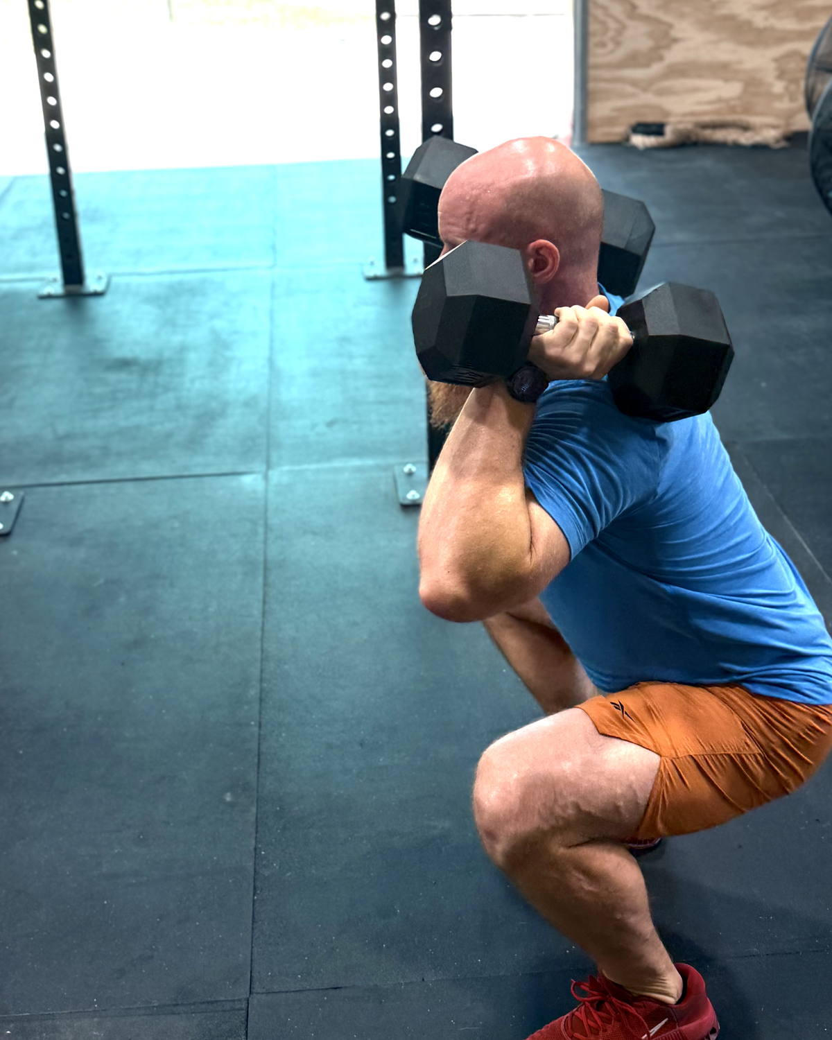 An athlete at the bottom of a dumbbell front squat, elbows driven high, alone on the gym floor.